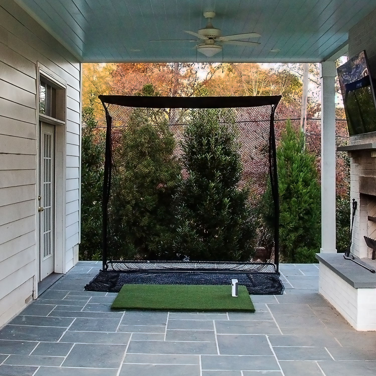 An outdoor backyard patio golf simulator setup featuring a black hitting net on a stone tile floor, complete with a green turf hitting mat and launch monitor positioning.